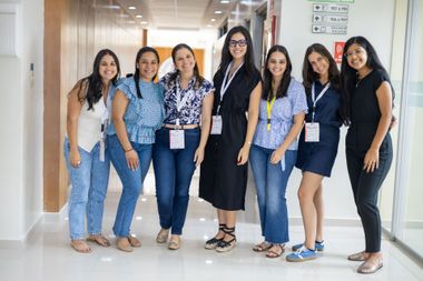 Seven women in a well-lit hallway smile at the camera. They wear casual clothes and lanyards.