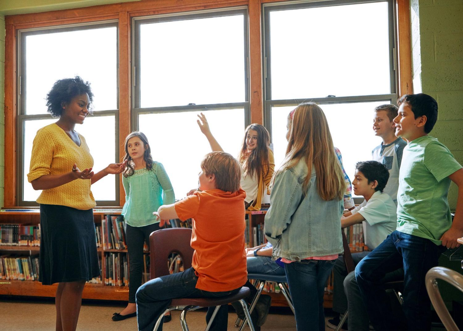 Teacher speaking to a group of students near a window in a library. One student raises their hand.