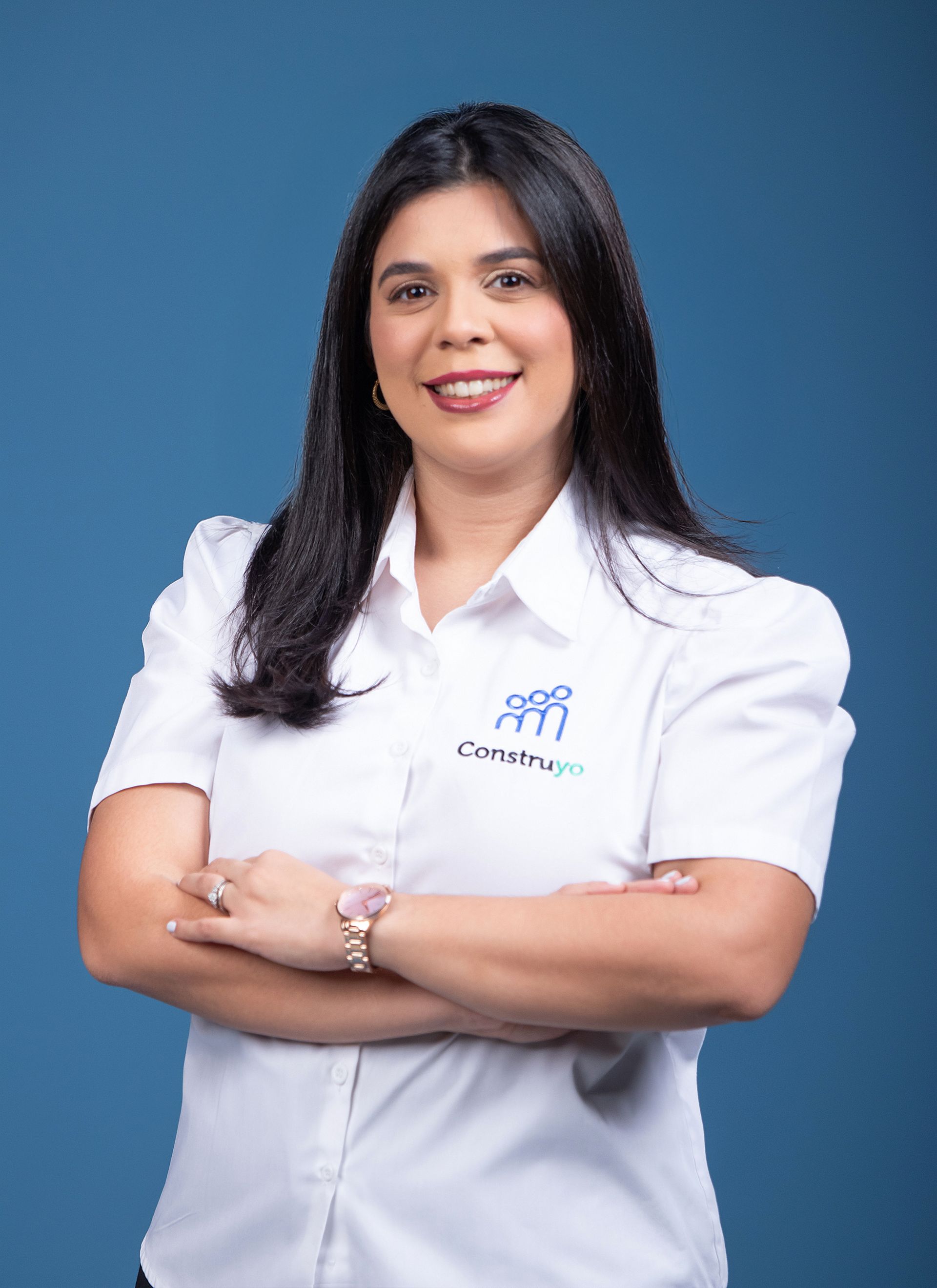 Woman with dark hair, crossed arms, wearing a white shirt with a logo, smiling, blue background.