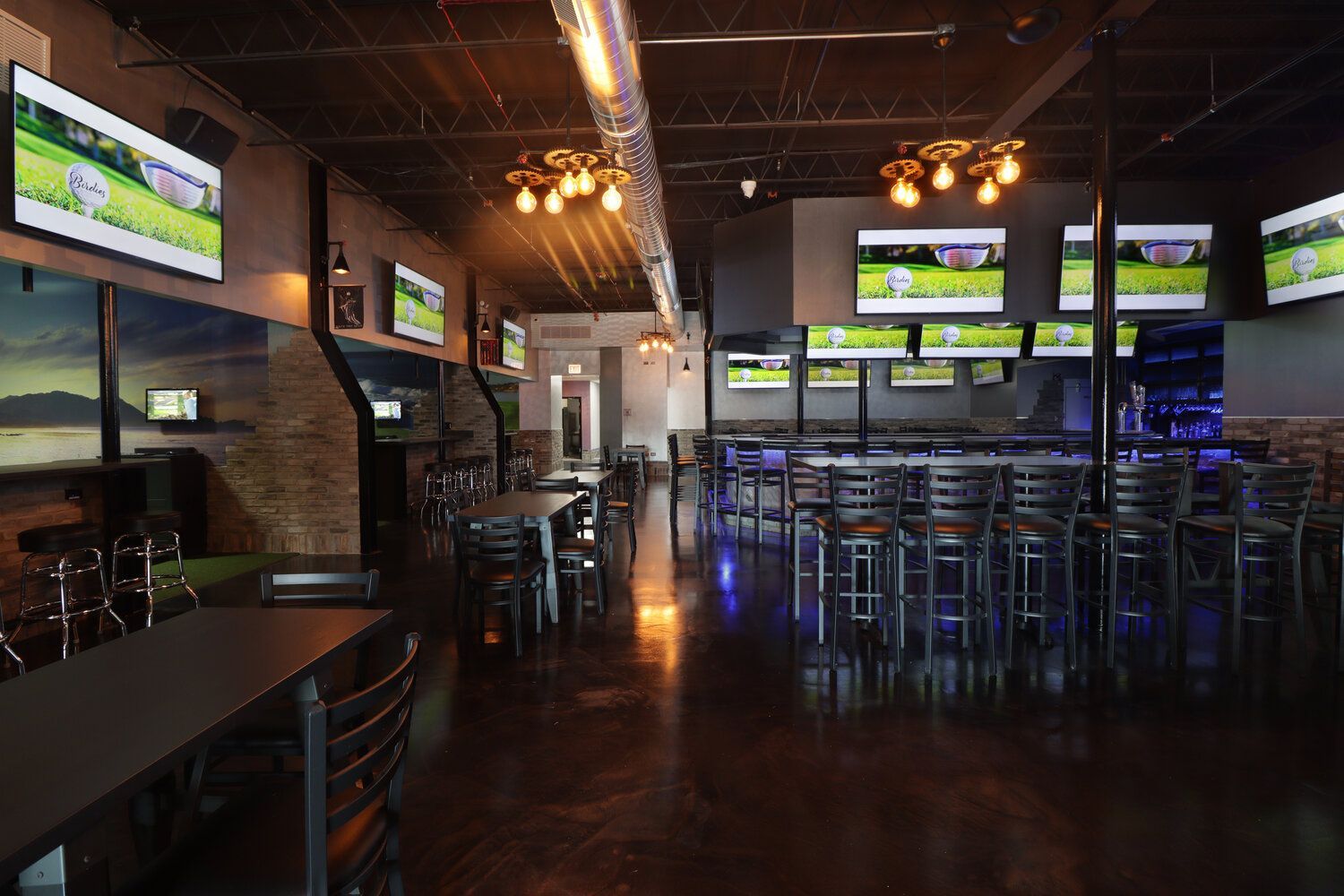 A restaurant with a lot of televisions on the wall and tables and chairs.