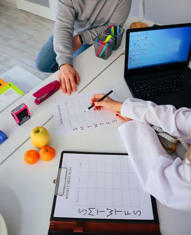 Two people working at a table with notebooks, laptop, markers, and fruit during a study session