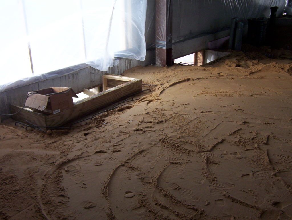 Sand-covered floor with a wooden tray and a covered piece of furniture by a wall.
