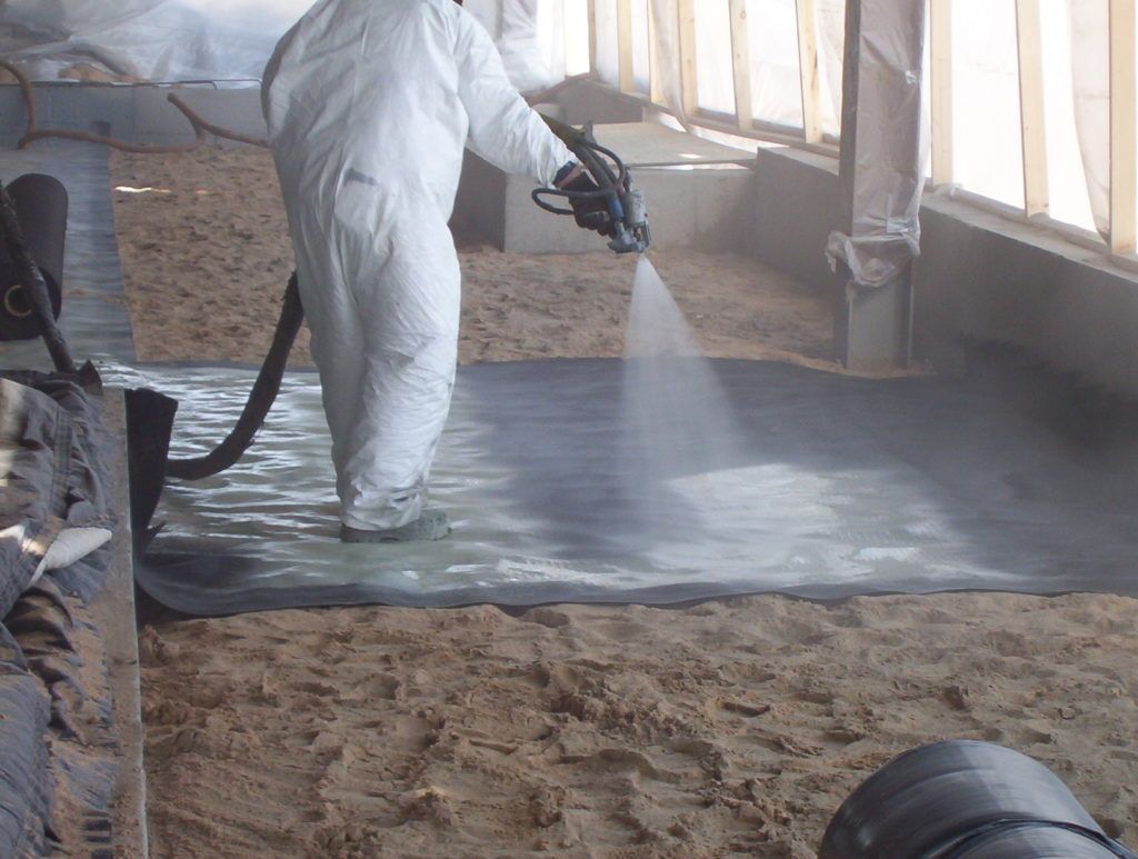 Worker in white protective suit spraying liquid onto a concrete floor in a dusty industrial room.