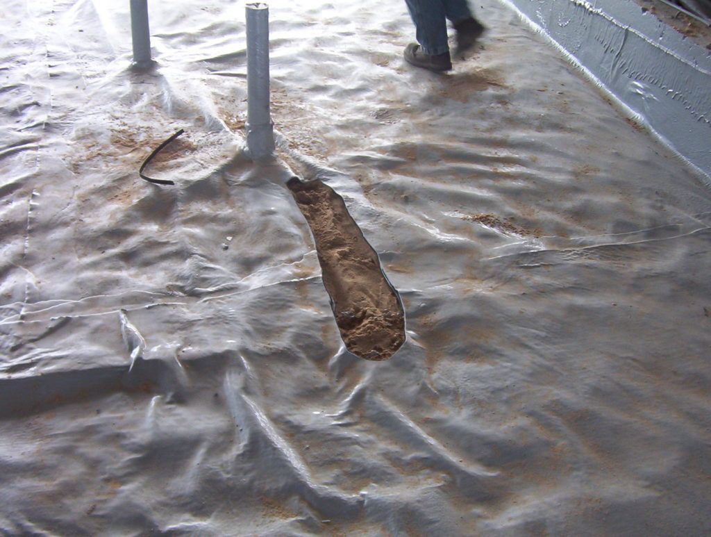 Mud-covered floor with broken concrete around two metal posts and a person’s feet in the background