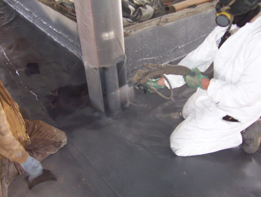 Worker in white protective suit kneels beside industrial machinery, handling a green tool on a dark floor.