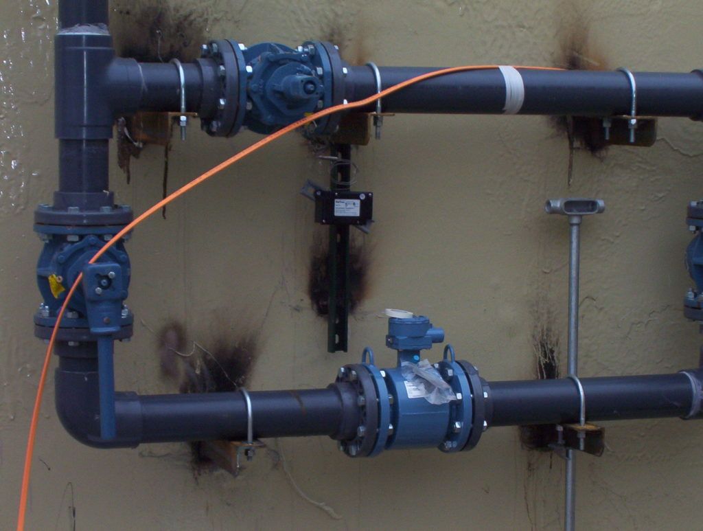 Blue industrial pipes and valves mounted on a beige wall, with an orange cable crossing the top pipe.
