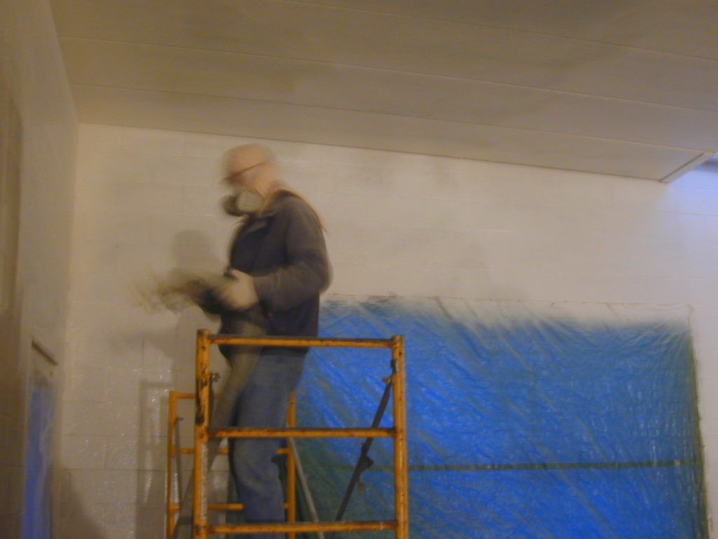 Worker on orange scaffold painting a white wall beside blue tarps inside a room