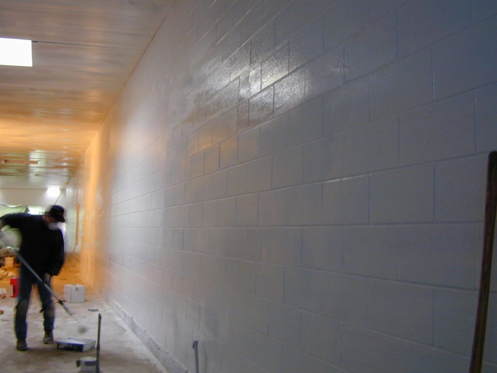Worker painting a cinderblock hallway wall with a roller and paint tray