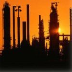 Silhouetted industrial refinery towers against an orange sunset sky
