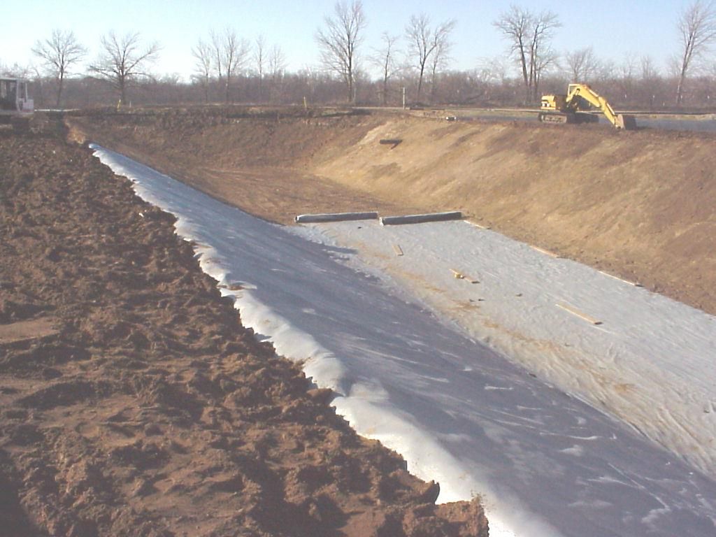 Excavation site with a sloped plastic-lined basin and a yellow excavator in the distance