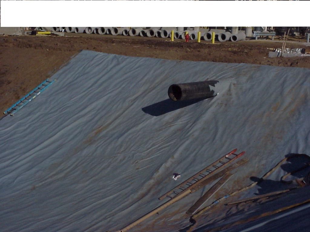Large gray tarp covering a sloped surface, with a black pipe and construction materials nearby.