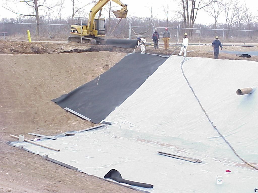 Workers spread black liner over a drainage pond with an excavator nearby on a construction site.