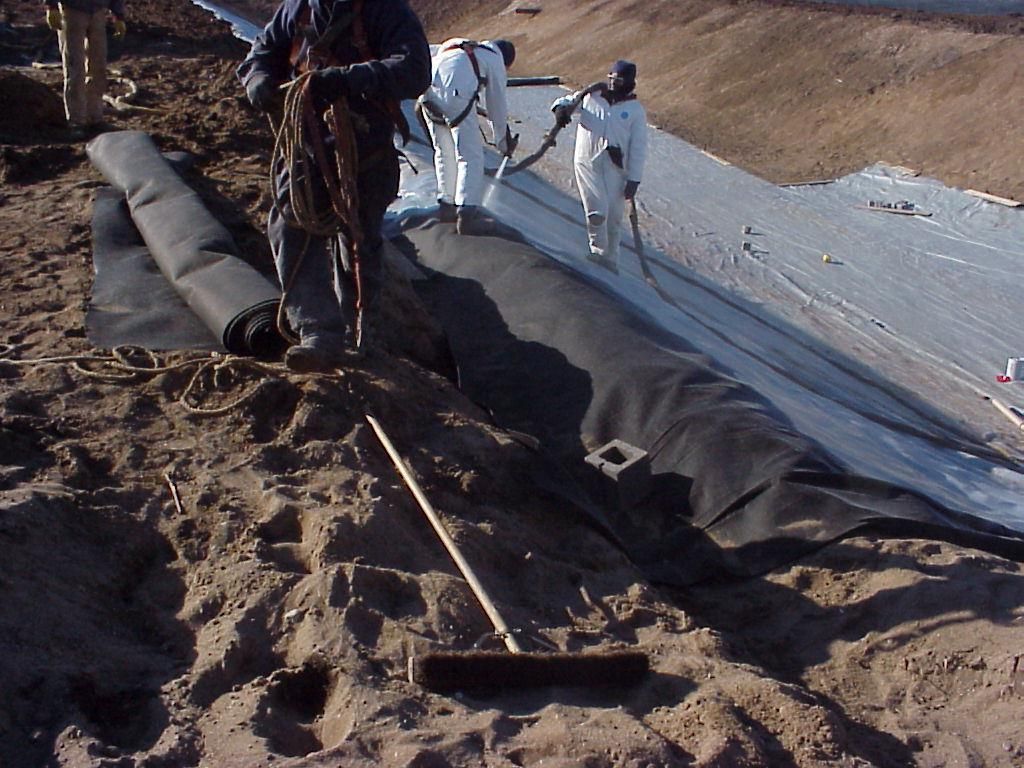 Workers installing black plastic sheeting on a dirt slope beside white ground cover