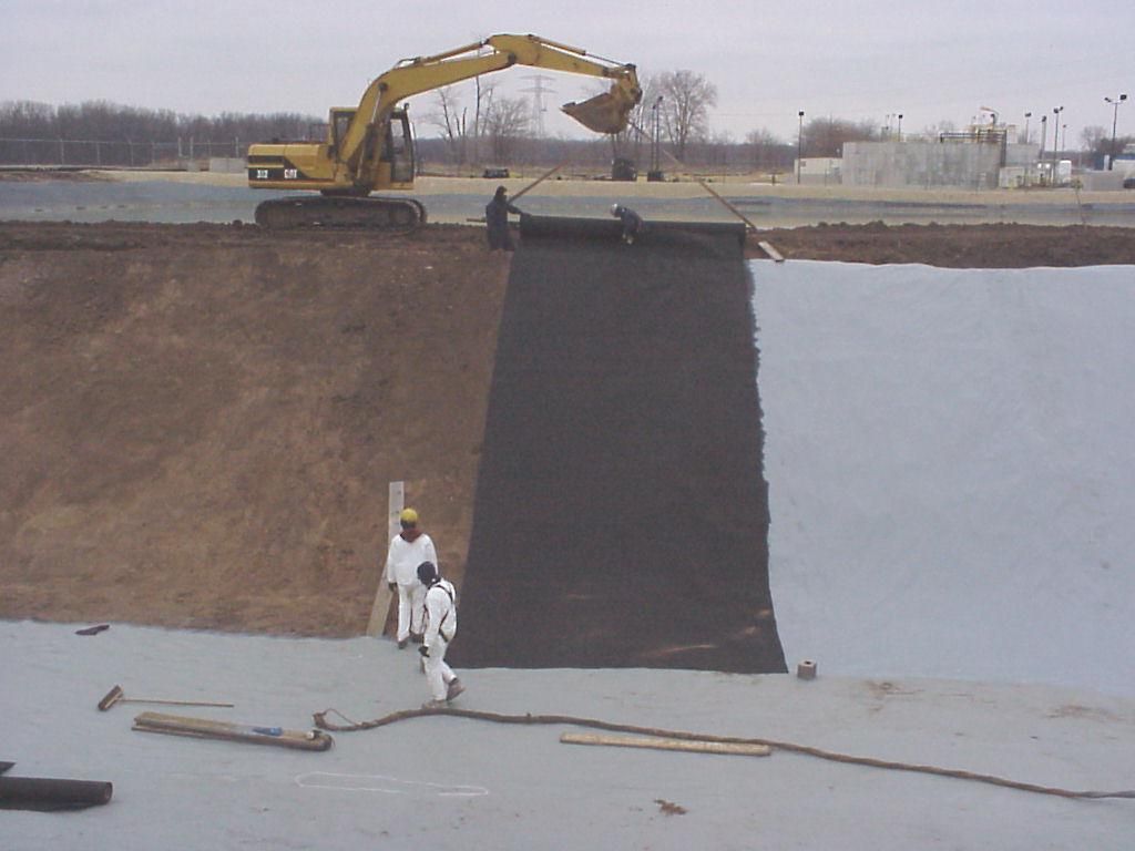Excavator working on a sloped embankment beside water, with two workers below holding a hose or cable.