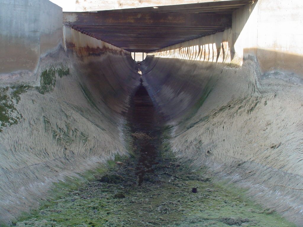 Concrete drainage ditch under a bridge, with shallow water and stained sloped walls.
