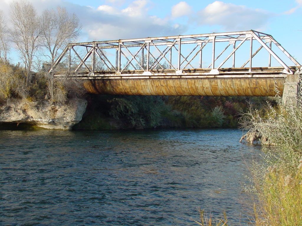 Rusty truss bridge over a blue river with rocky banks and trees under a partly cloudy sky