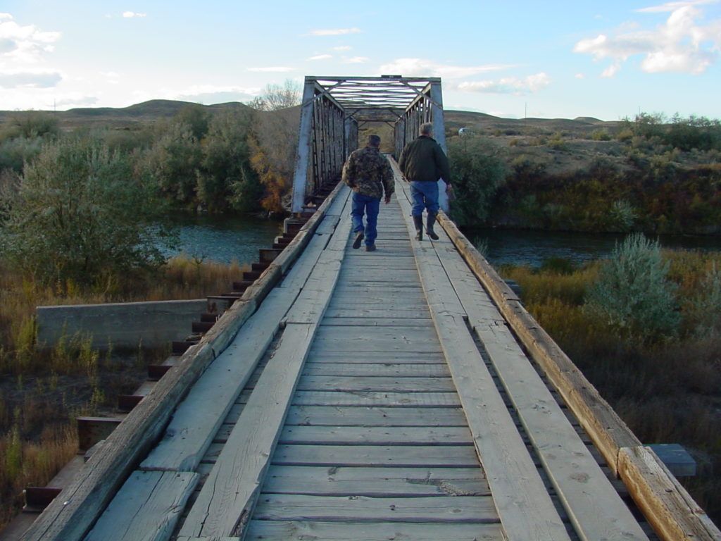 Two people walking across a narrow wooden bridge over a marsh at dusk