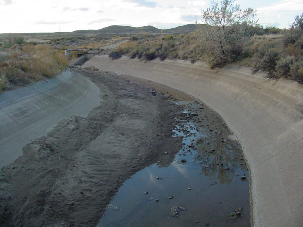 Dry concrete canal with shallow muddy water winding through a desert landscape under a cloudy sky