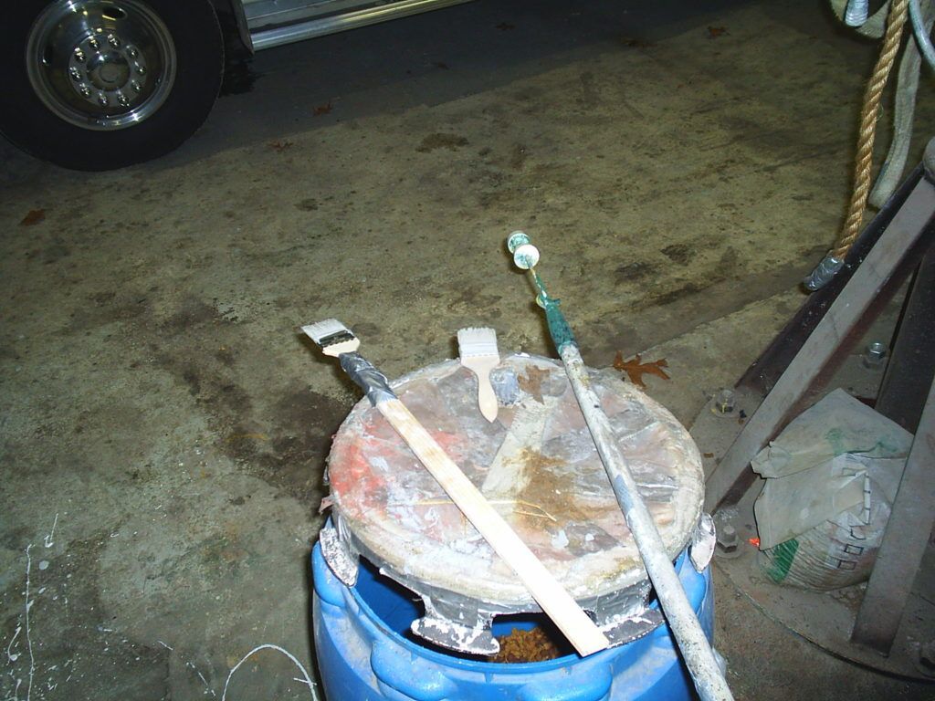 Rusty metal drum with a lid and pipes/rods on top, sitting on a concrete floor in a garage.