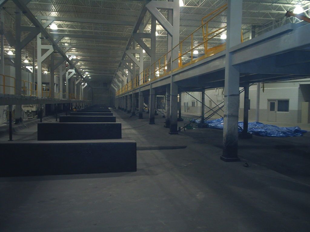 Large industrial warehouse interior with metal beams, raised platform, and stacked dark materials on the floor