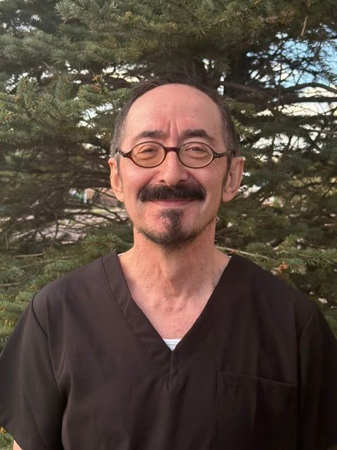 Smiling person with glasses and a mustache, wearing a dark shirt outdoors with trees behind them Smiling person with glasses and a mustache, wearing a dark shirt outdoors with trees behind them