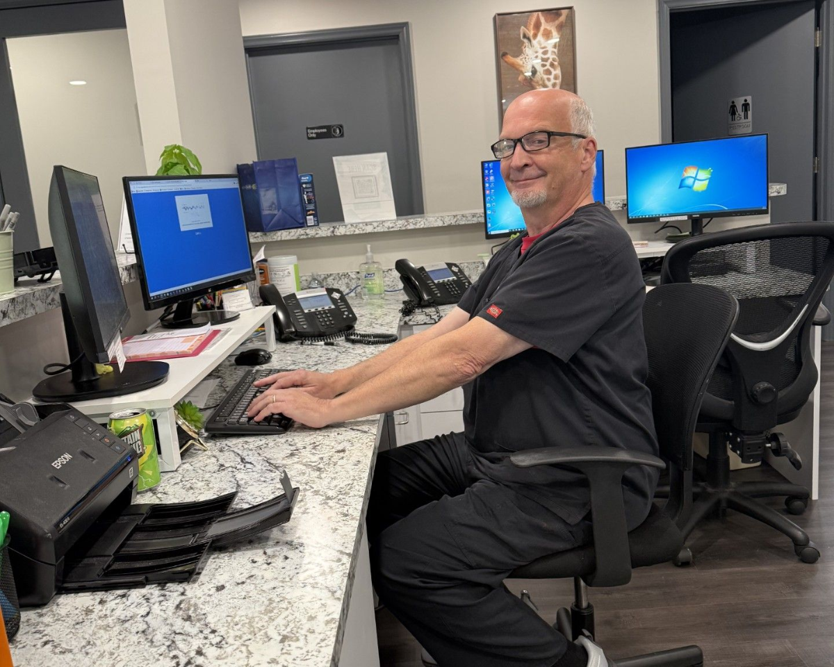 Man in scrubs at a computer, smiling, at a reception desk with two monitors and a printer.