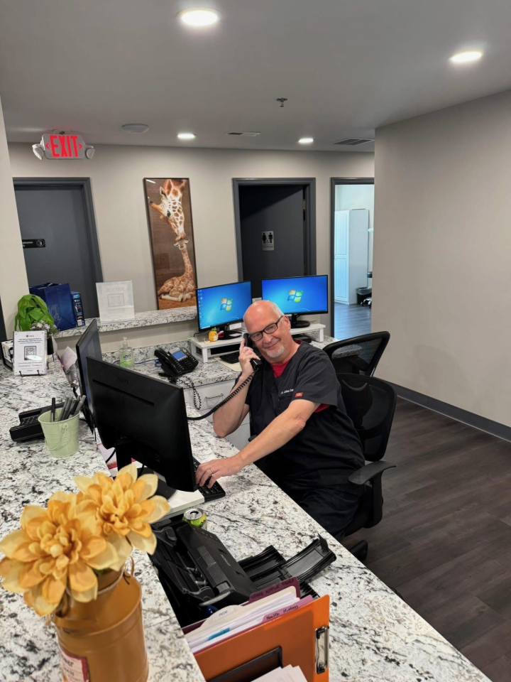 Man in scrubs at a reception desk, talking on the phone and smiling. Office setting with two monitors.