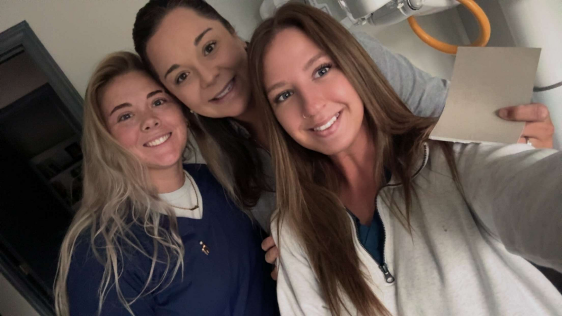 Three smiling women taking a selfie; blonde, brunette, and dark-haired, indoors.