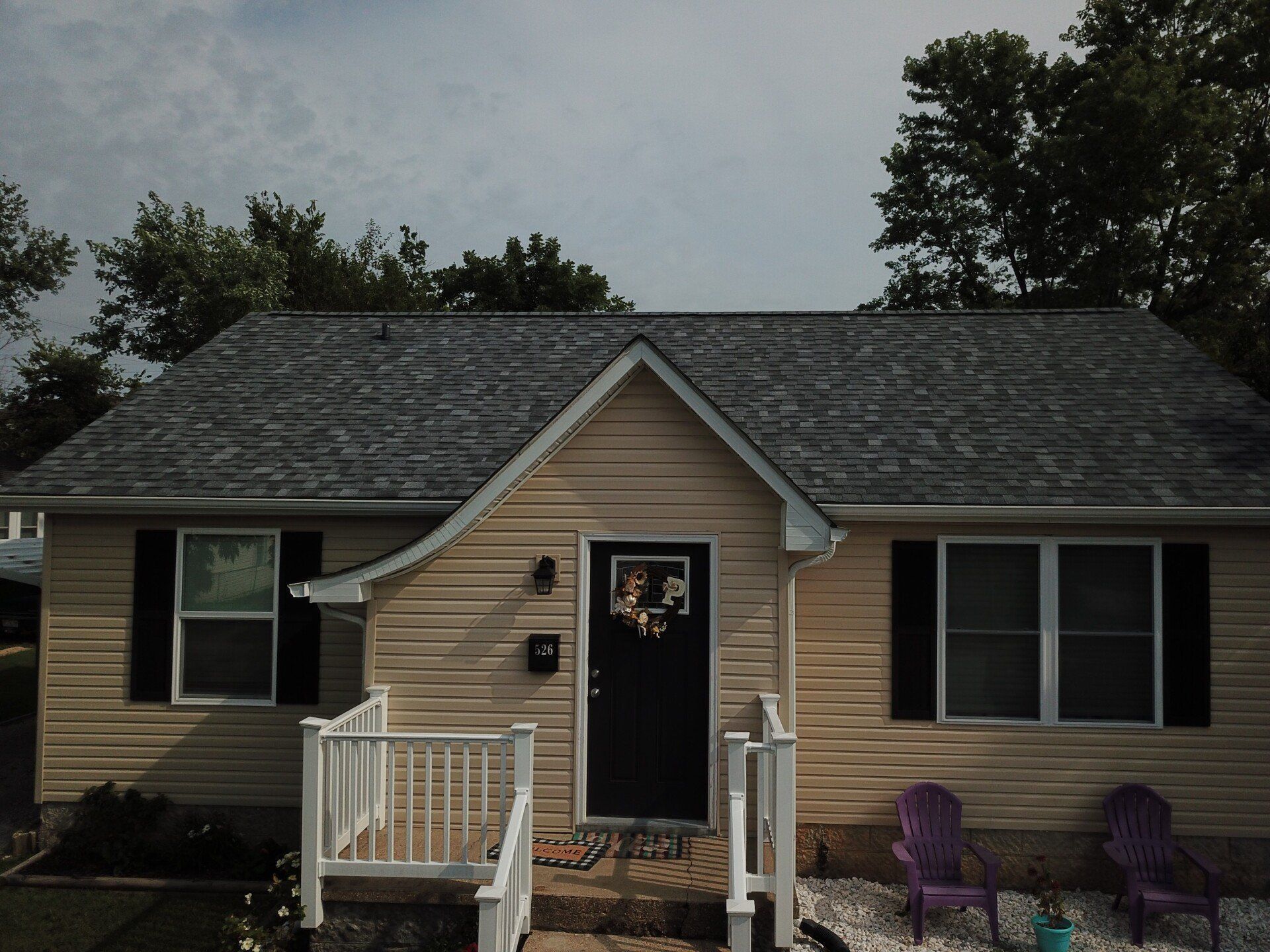 Small beige house with black shutters, front porch, and white railing, seen from the front.
