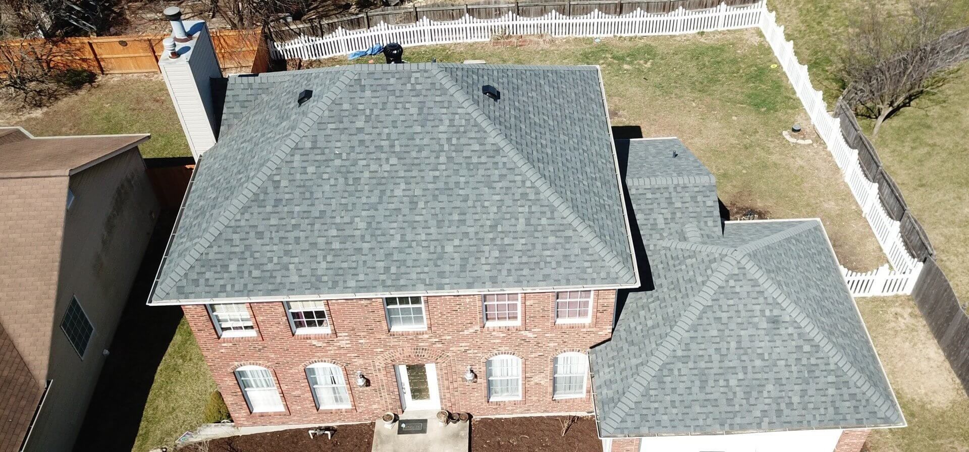 Aerial view of a brick house with gray shingle roofs and a fenced backyard.