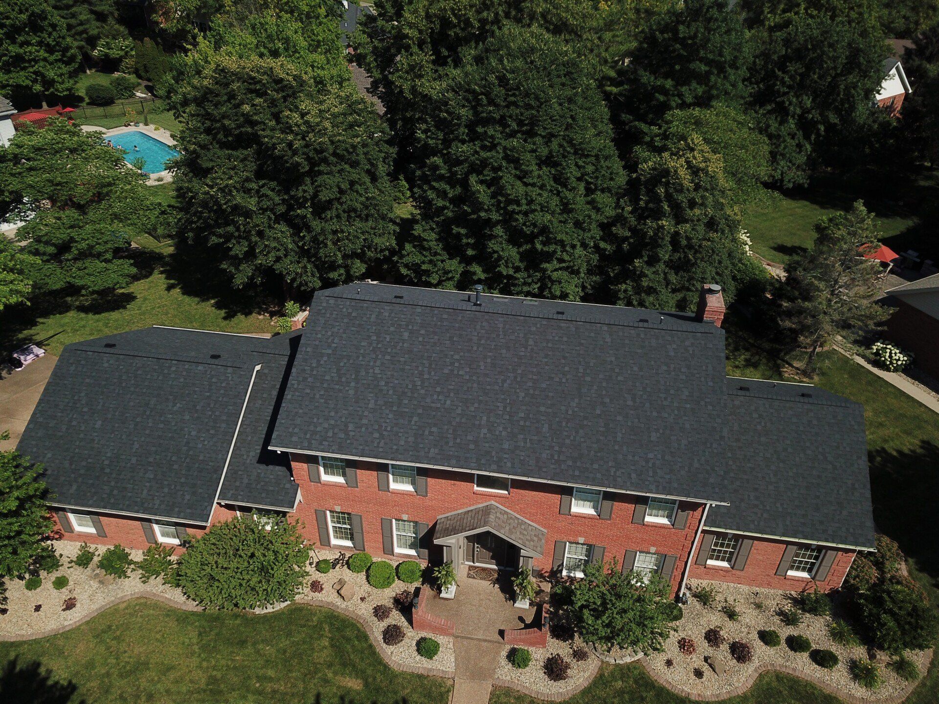 Aerial view of a red brick house with a dark roof, surrounded by trees and a landscaped front yard.