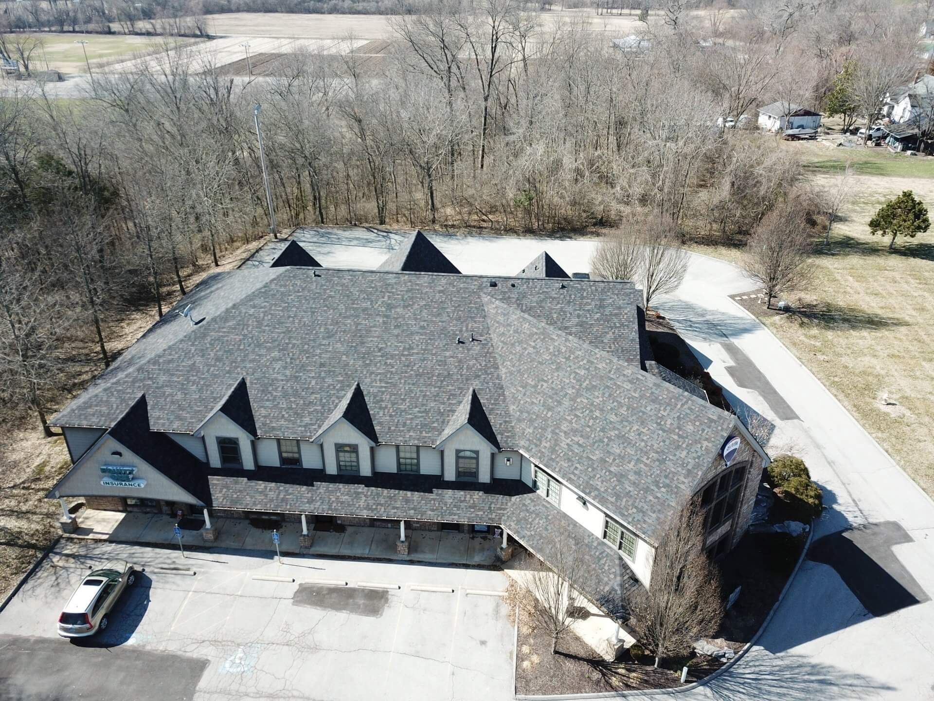 Aerial view of a large gray-roofed building with parking lot, surrounded by trees and open land