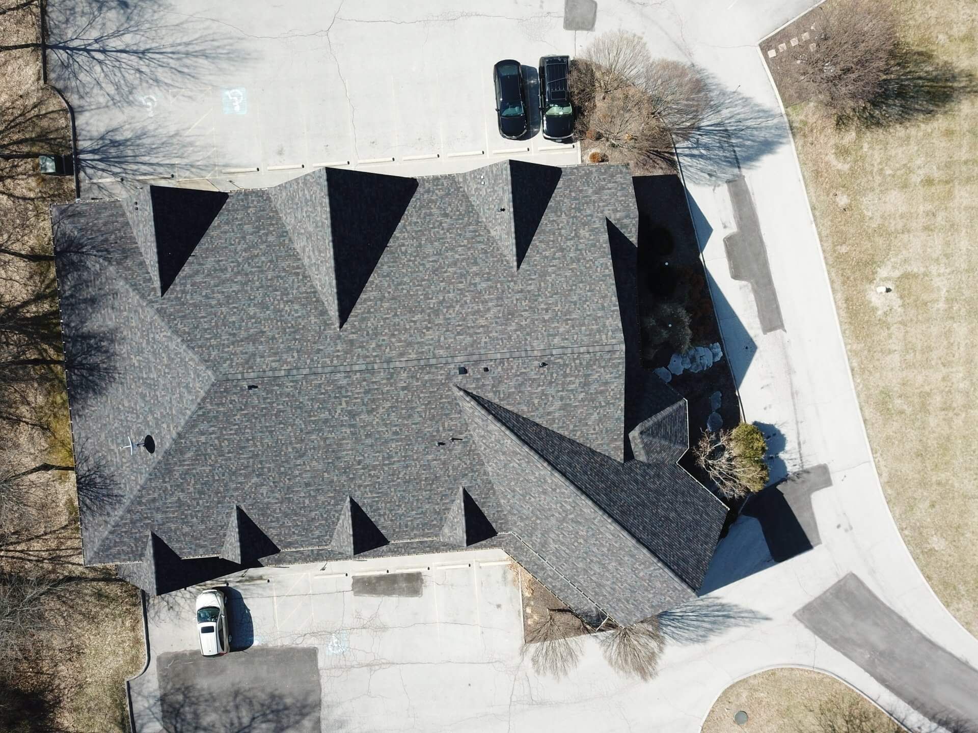 Aerial view of a gray-roofed house with driveway, parked cars, and surrounding trees.