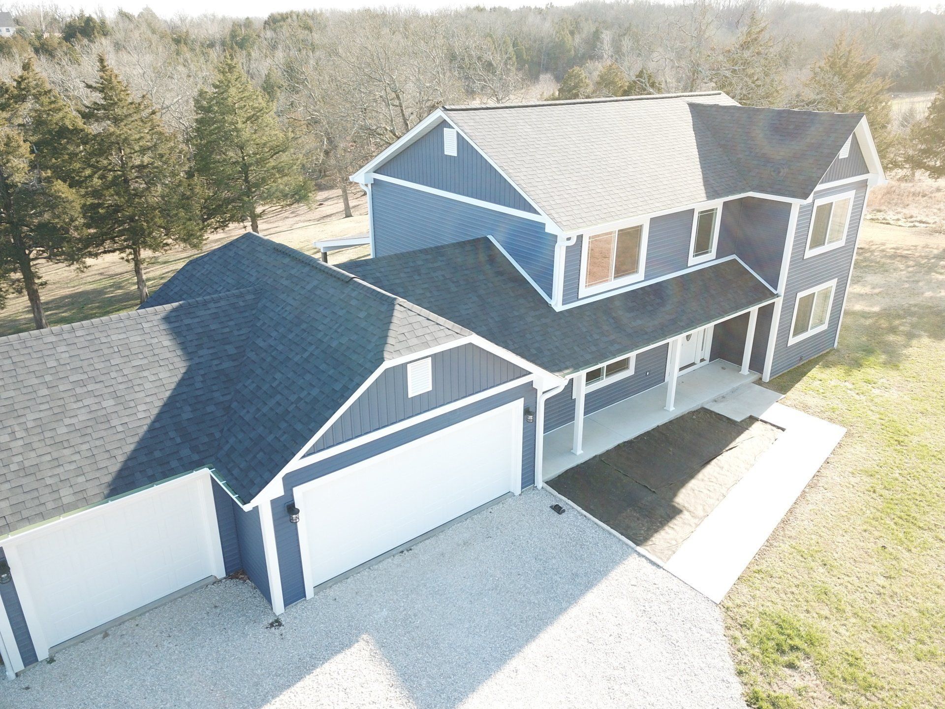Aerial view of a two-story blue house with a double garage and driveway surrounded by trees.
