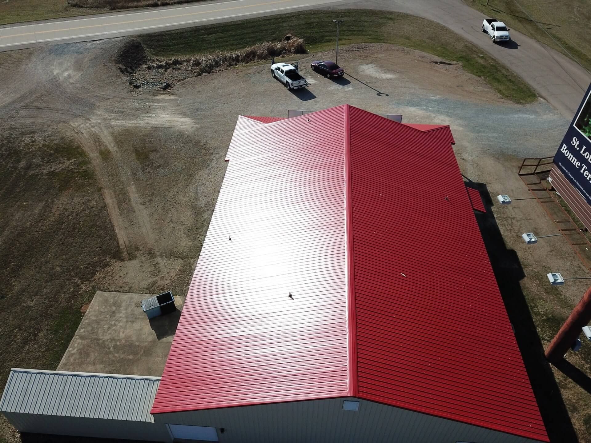 Aerial view of a red-roofed building with a white glare, dirt lot, and parked vehicles nearby.