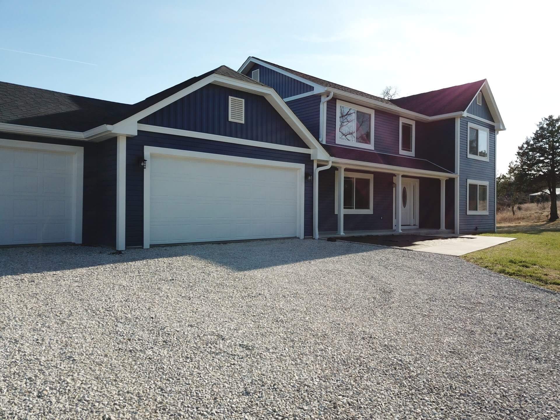 Blue two-story house with white garage doors and gravel driveway under a clear sky