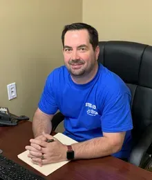 A man in a blue shirt is sitting at a desk with his hands folded.