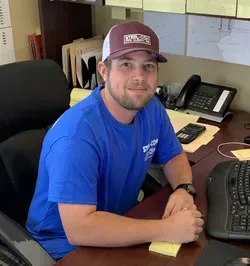 A man in a blue shirt and hat is sitting at a desk.