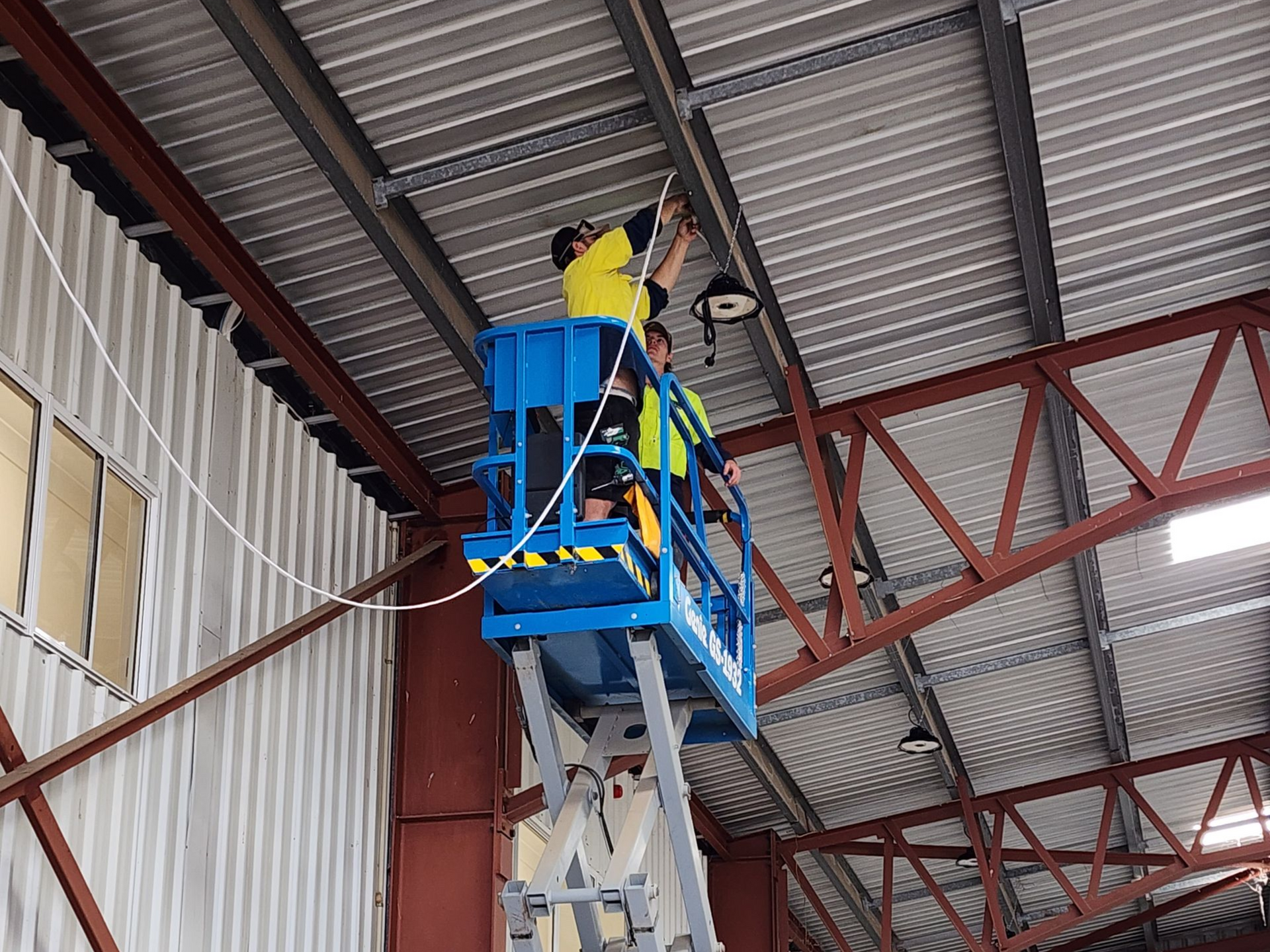 Worker on a blue lift painting a metal ceiling inside an industrial building — Bes-Elect Electrical in Taminda, NSW