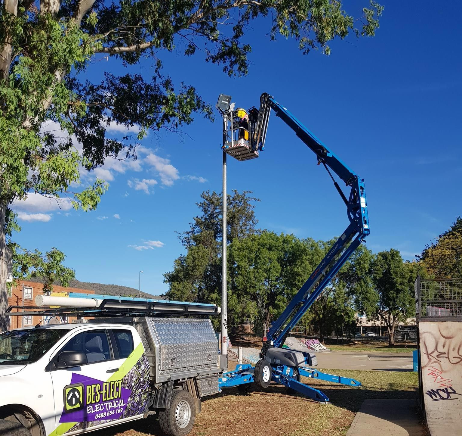 Blue Cherry Picker Lifts Worker Beside a Utility Pole on A Sunny Street — Bes-Elect Electrical in Taminda, NSW