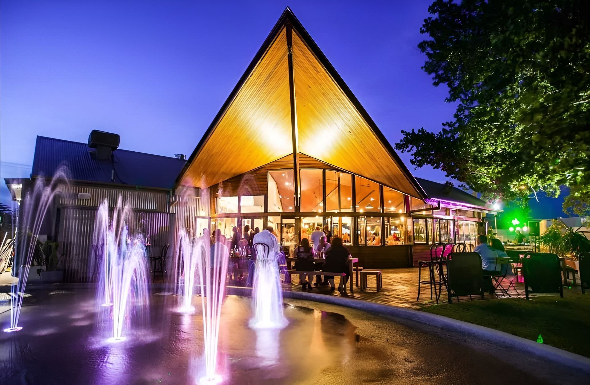 Lit Fountain Outside a Glass Pavilion at Dusk with People Dining Inside — Bes-Elect Electrical in Taminda, NSW