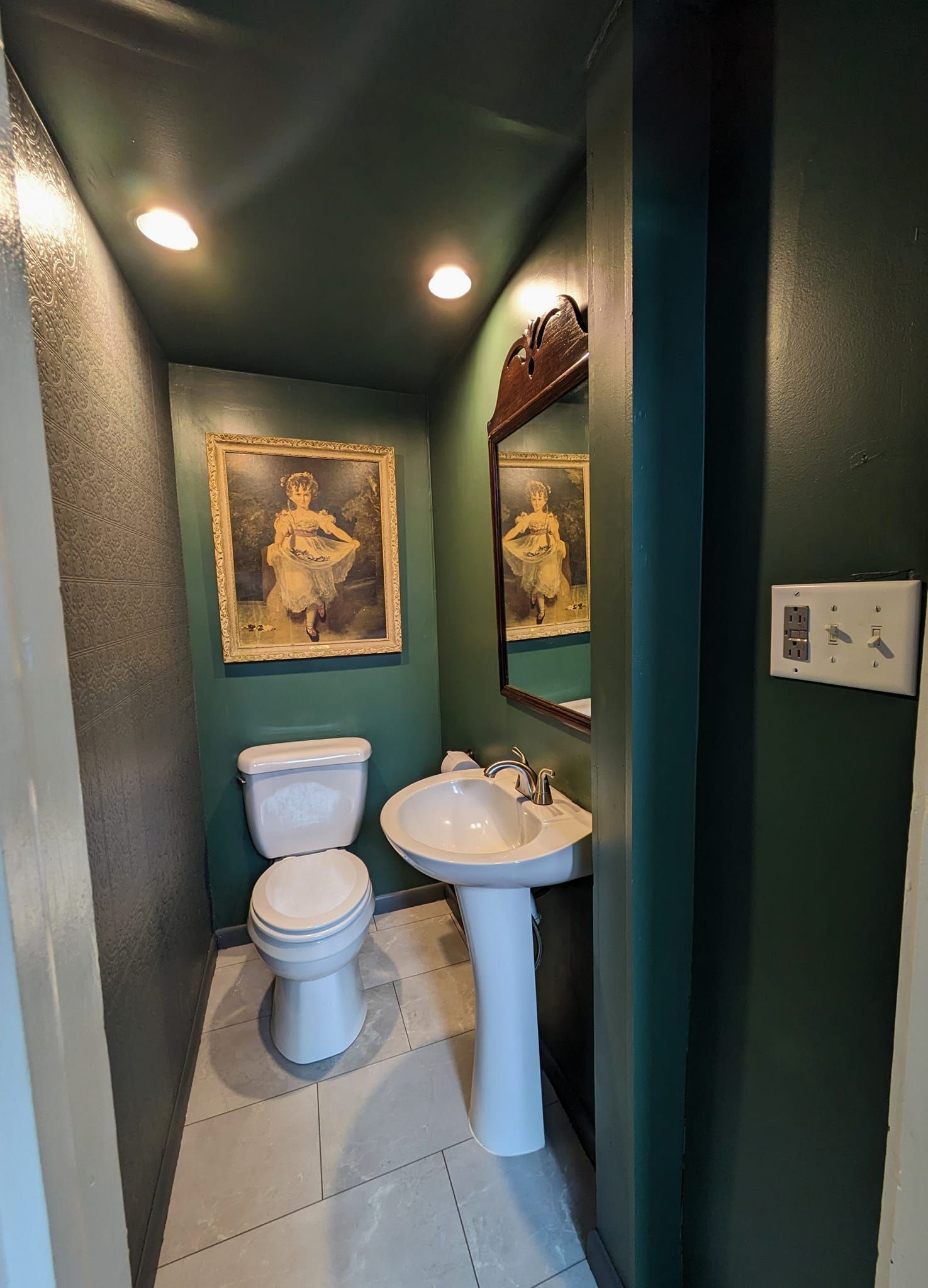 Narrow green bathroom with toilet, pedestal sink, and framed wall art under ceiling lights