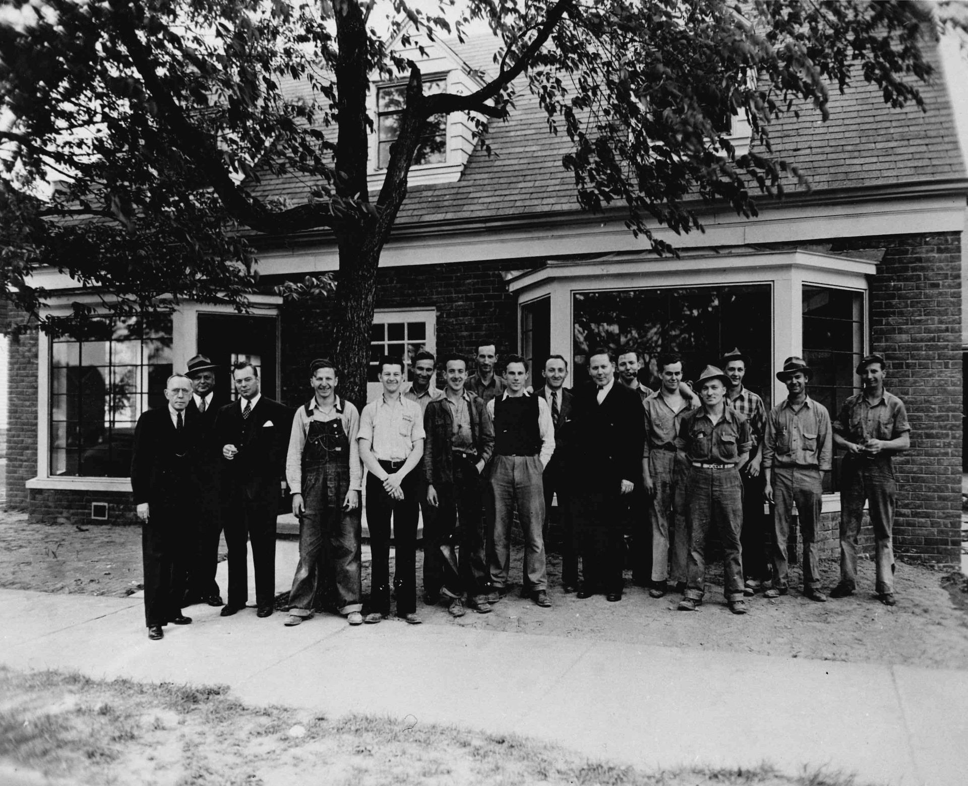Group of people standing outside a brick house with a porch, posing for a black-and-white photo.