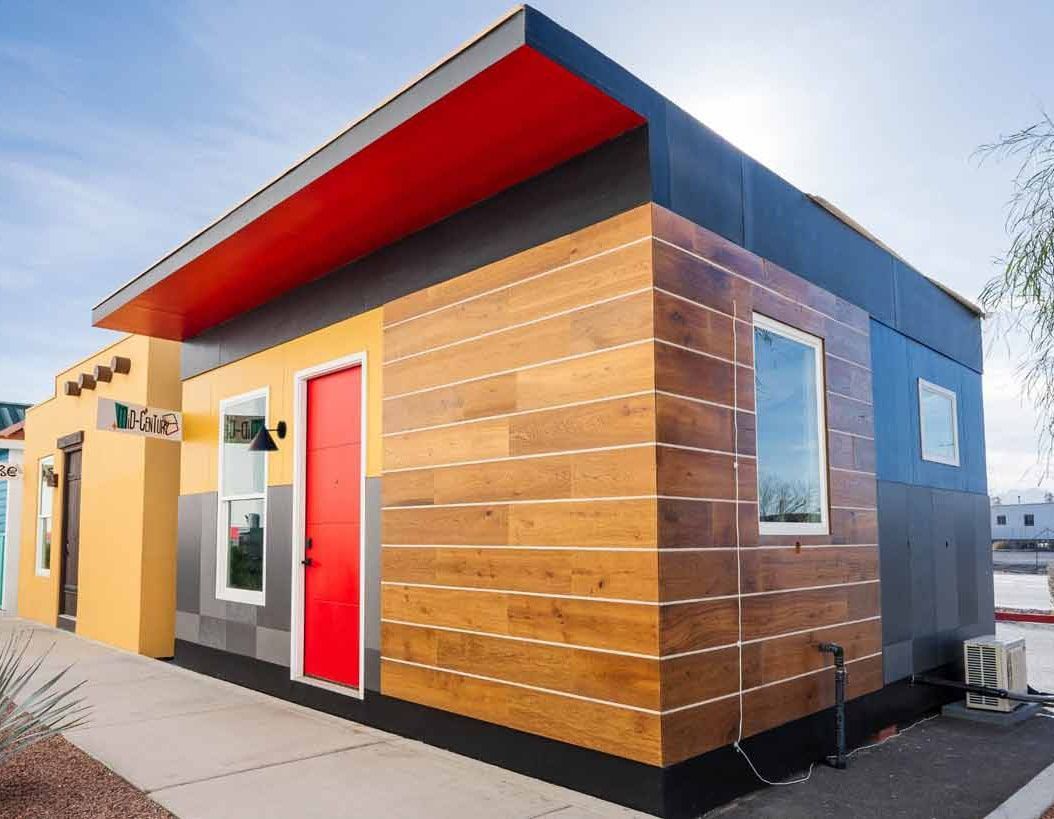 Modern small building with wood siding, red roof overhang, and colorful doors under a clear sky