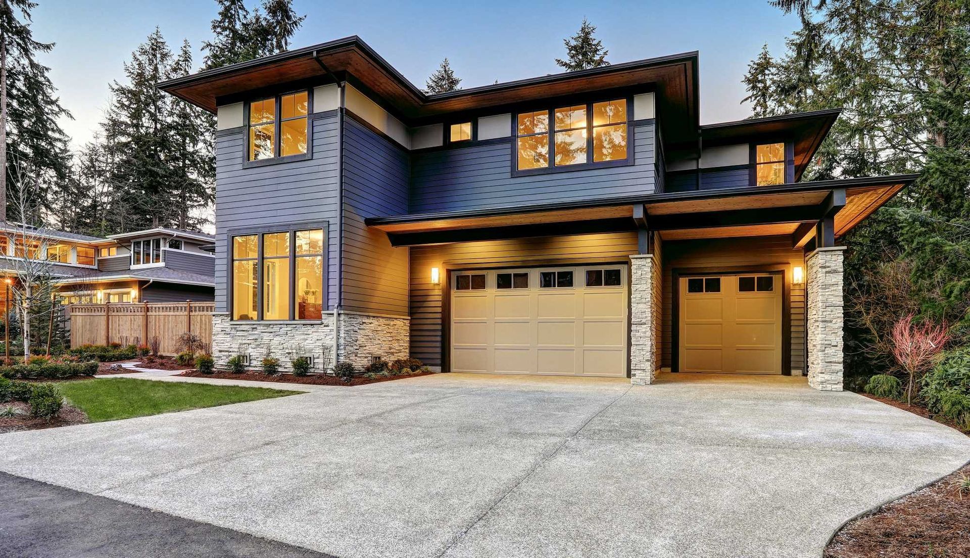 Modern two-story house with blue siding, warm-lit windows, and a three-car garage at dusk