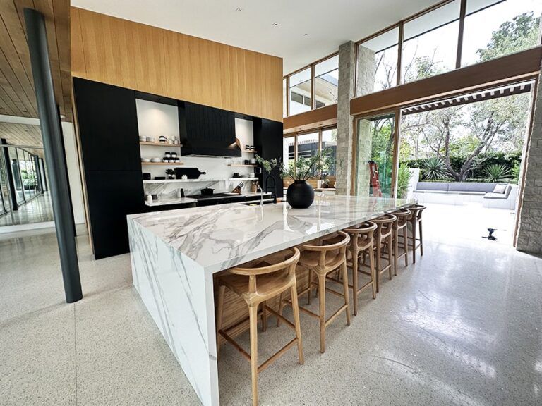 Modern kitchen with marble island, wooden stools, and floor-to-ceiling windows opening to a patio