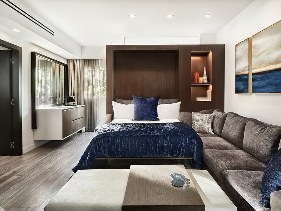 Modern bedroom with a dark wood bed, blue bedding, gray sofa, and large windows.