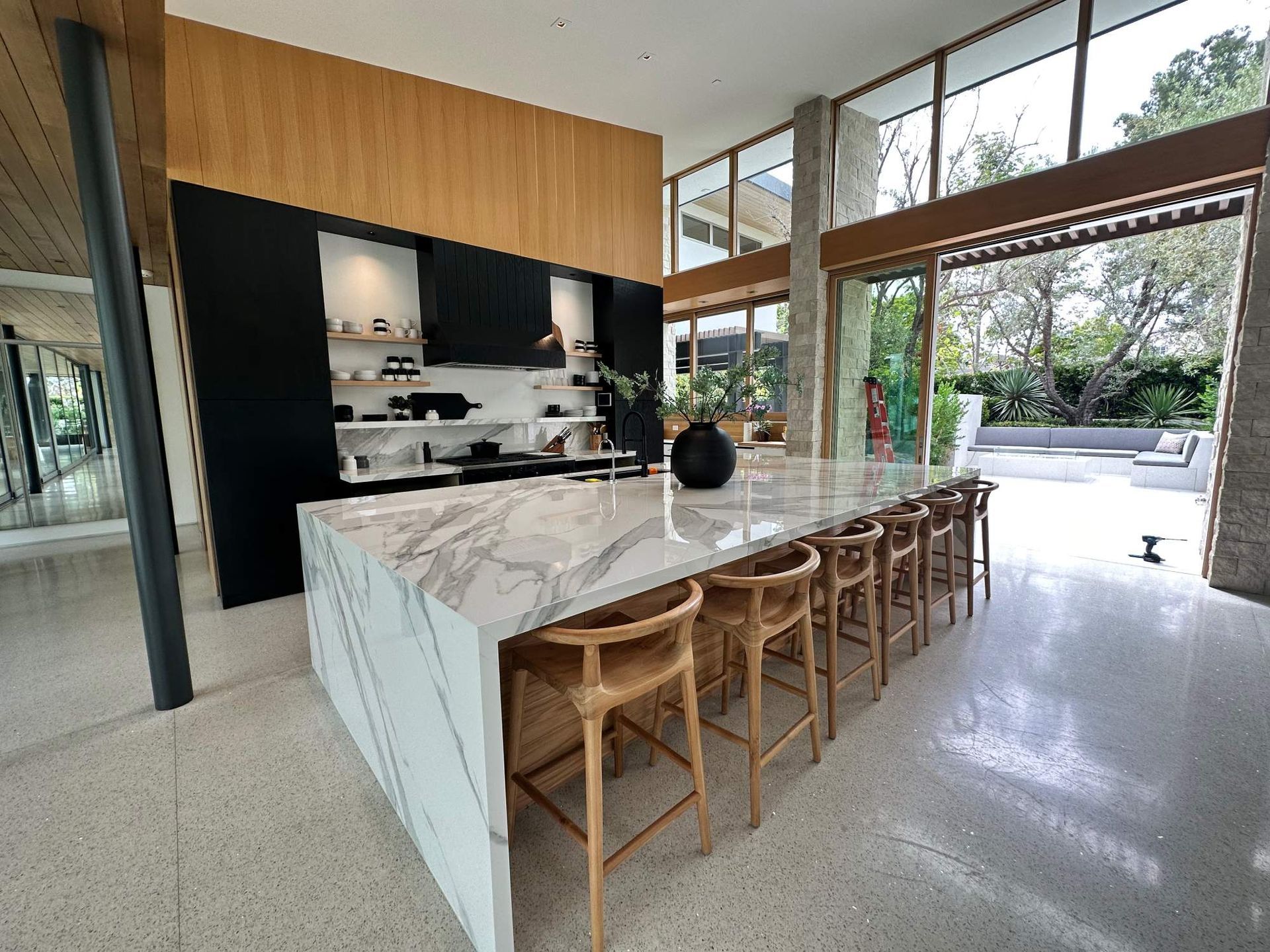 Modern kitchen with marble island, wooden stools, and large glass walls opening to a patio and garden