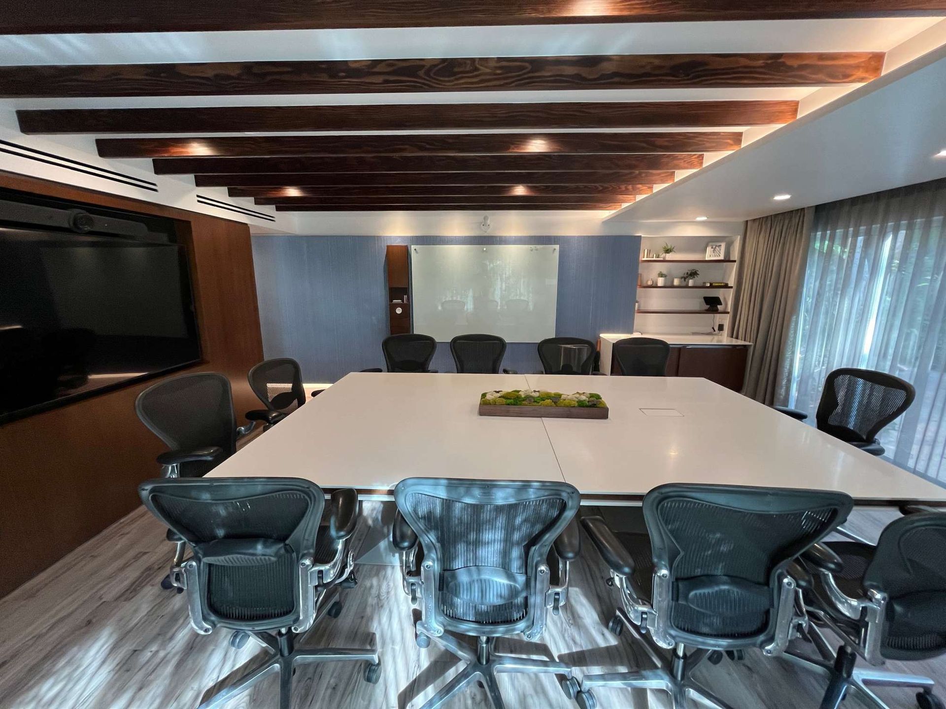 Modern conference room with large white table, black chairs, wood beam ceiling, and wall-mounted TV