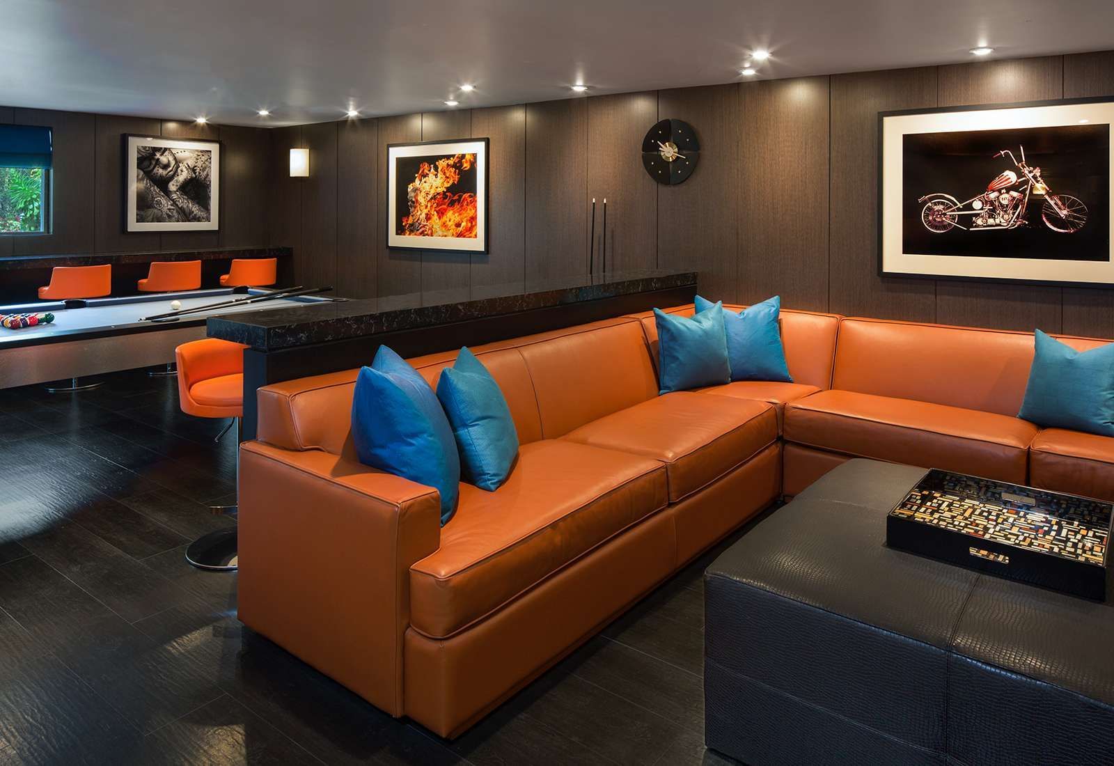 Modern lounge with tan sectional sofas, blue pillows, dark walls, framed art, and recessed lighting.